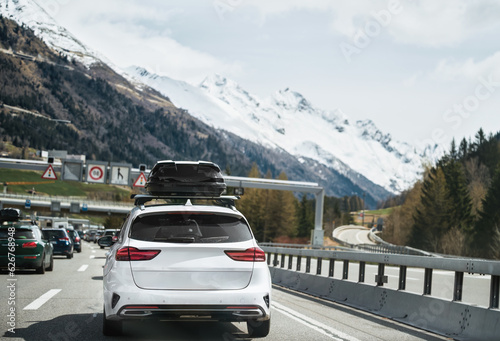 Modern Family Car Adventures in Switzerland. View from the back of a black plastic luggage box on a car roof. Alpine Road Trip Getaway. White family wagon car © AlexGo