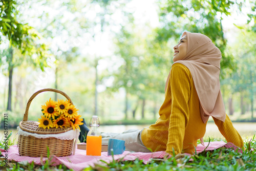 Happy Asian muslim woman sitting on the grass field in garden. Muslim woman having a picnic in garden.