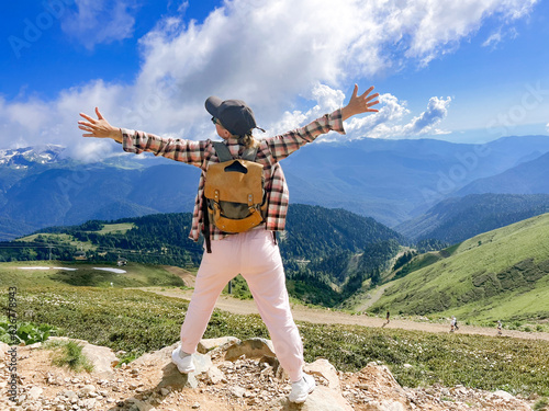 young stylish woman hiking alone with backpack enjoying beauty of mountains with hands raised up
