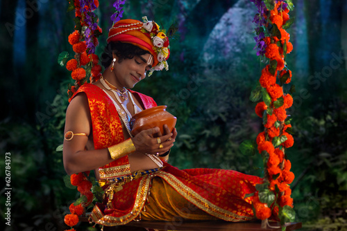 Young man dressed up as Lord Krishna and sitting on a swing with a butter pot on the occasion of Janmashtami photo