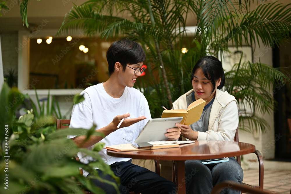 Two happy Asian college students are sitting at a cafe in an outdoor area, discussing work
