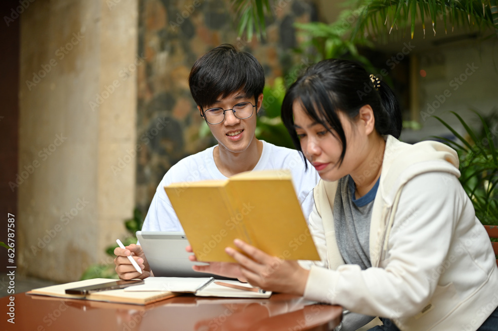 A focused Asian female college student is reading a book while her male friend is using a tablet A focused Asian female college student is reading a book while her male friend is using a tablet