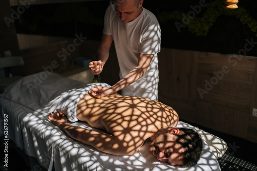 Professional manual therapist massaging man back with oil during relaxing or revitalizing procedure at Thai beauty spa salon. Guy client lying on couch enjoying body and skin care in wellness center..