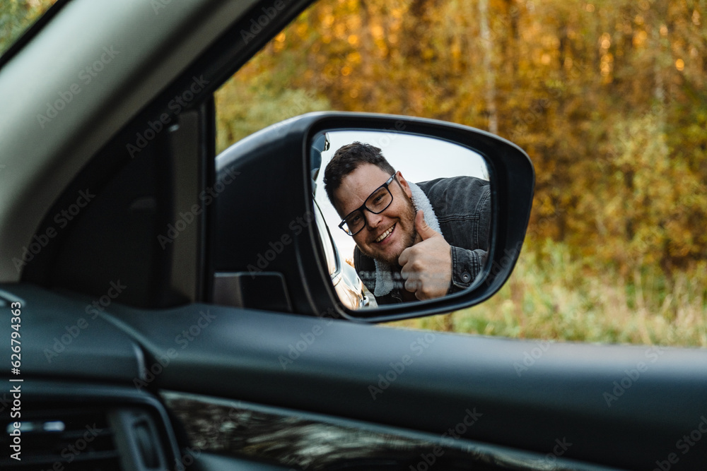 A smiling man in the side mirror of the car shows a big stick up behind the autumn forest