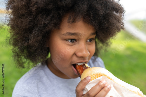 Dark skinned female child eating junk food with pleasure in park while sitting on green grass.