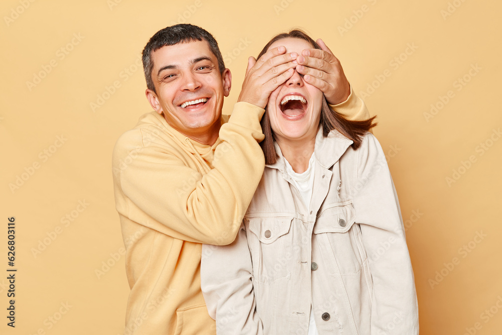 Romantic man covering girlfriend's eyes while standing together, loving couple posing isolated over beige background, having fun, laughing. Romantic man covering girlfriend's eyes while standing together, loving couple posing isolated over beige background, having fun, laughing.