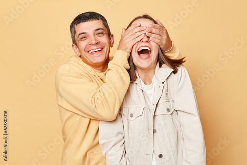 Romantic man covering girlfriend's eyes while standing together, loving couple posing isolated over beige background, having fun, laughing.