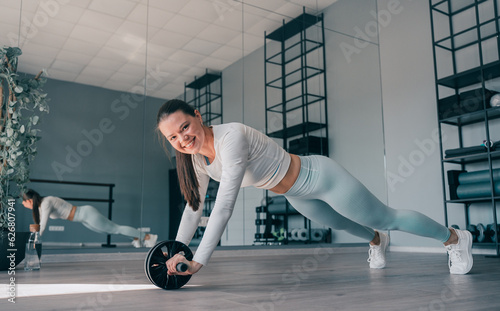 Brunette cheerful young woman in sportswear at abdominal exercise, rollouts. Fit caucasian girl improving body endurance, Happy fitness model at workout, looks air camera toothy smiles.