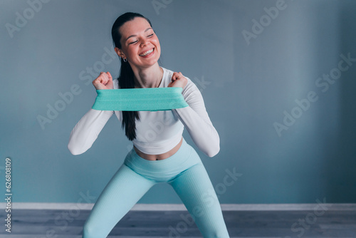 Cheerful caucasian young woman in sportswear stretcing rubber tape making squats at sport club. Brunette American girl at endurance exercise broad smiles against grey wall. Healthy people, fitness. photo