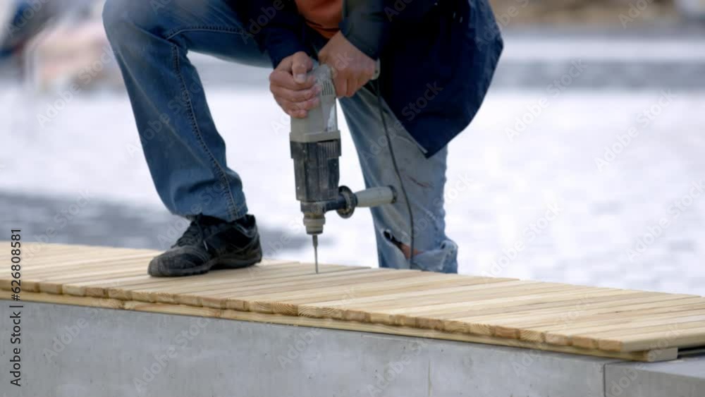Employee uses drill to create holes in bench boards in park. Worker fastens wooden planks together and forms bench during renovation works Employee uses drill to create holes in bench boards in park. Worker fastens wooden planks together and forms bench during renovation works