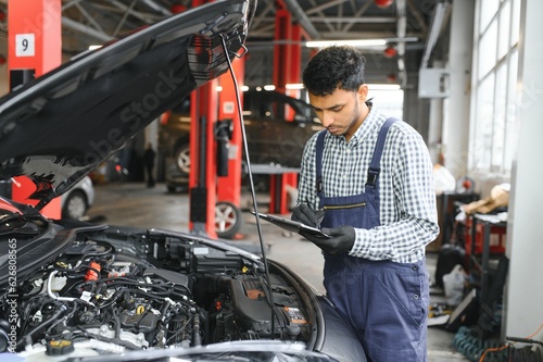 Indian car mechanic standing and working in service station. Car specialists examining the lifted car. Professional repairmen wearing mechanic uniform in blue color.