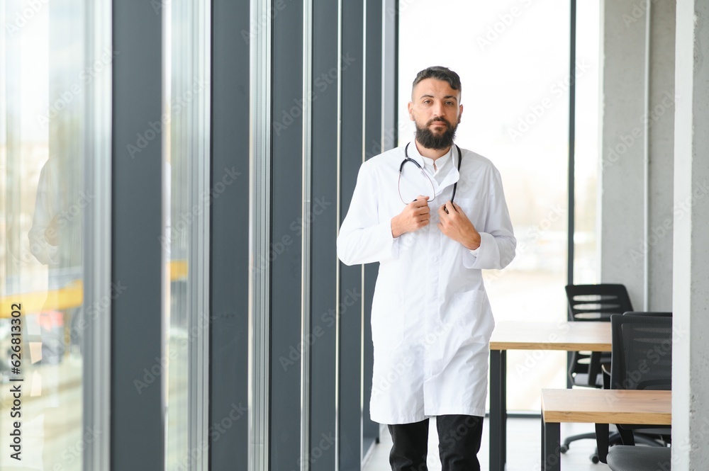 Young 30-aged handsome pleasant Arabic male doctor in white coat, posing at camera indoors. Young 30-aged handsome pleasant Arabic male doctor in white coat, posing at camera indoors.