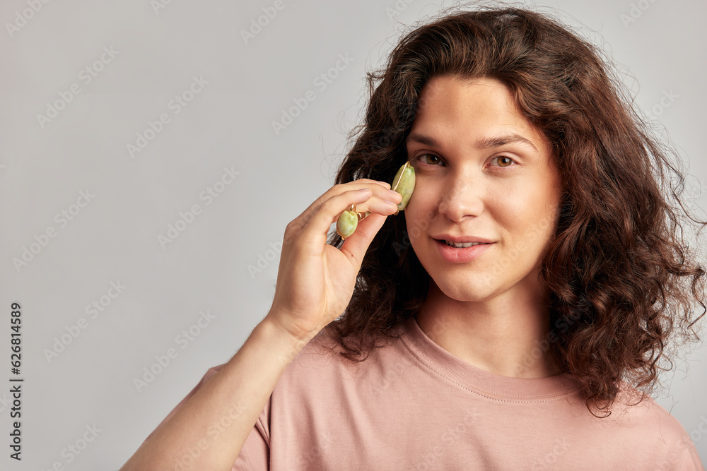 Young handsome long haired guy using jade de-puffing face roller to keep his skin soft and smooth. Caucasian man with brown hair maintaining his natural beauty with skincare routine. Men's self-care. Young handsome long haired guy using jade de-puffing face roller to keep his skin soft and smooth. Caucasian man with brown hair maintaining his natural beauty with skincare routine. Men's self-care.