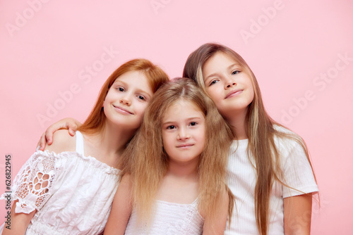 Three beautiful little girls, children in white clothes standing closely to each other, looking at camera against pink studio background. Concept of skincare, childhood, health, beauty products, ad