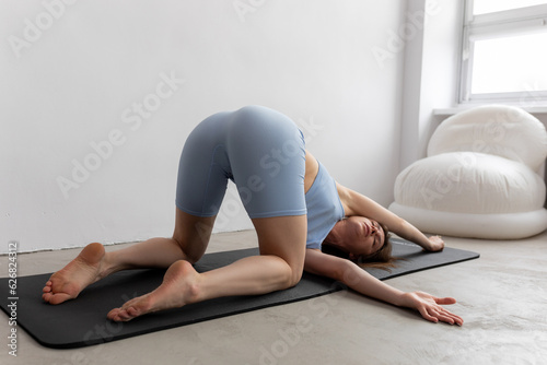 Portrait of young woman doing exercise, turning body, stretching, practising yoga on black mat on floor near armchair.