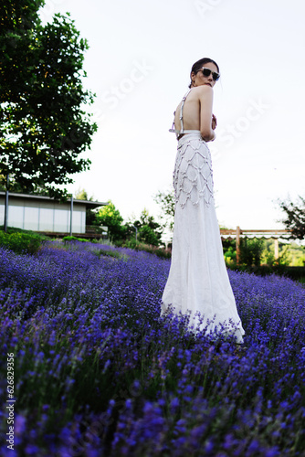 Beautiful young brunette woman in white dress and sunglasses posing in a lavender field at summer evening. Beauty concept