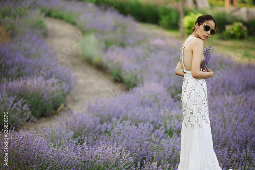 Beautiful young brunette woman in white dress and sunglasses posing in a lavender field at summer evening. Beauty concept