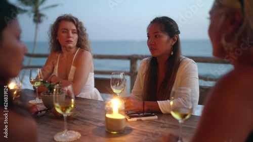 Diverse female friends drink wine in rooftop sea view restaurant at sunset. Multiracial women talking and smiling with candles at table in a tropical cafe at dusk. Young multiethnic women dining.
