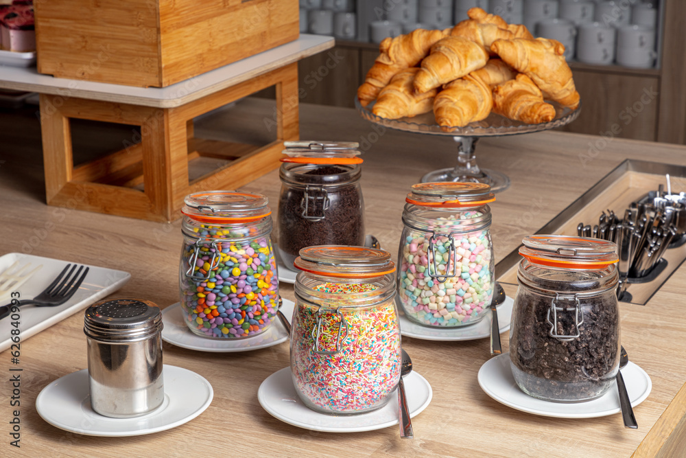 hotel breakfast buffet. a wooden table topped with jars filled with food and sprinkles and croissants in the background hotel breakfast buffet. a wooden table topped with jars filled with food and sprinkles and croissants in the background