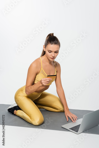 woman in sportswear using laptop and holding credit card while sitting on fitness mat, white background
