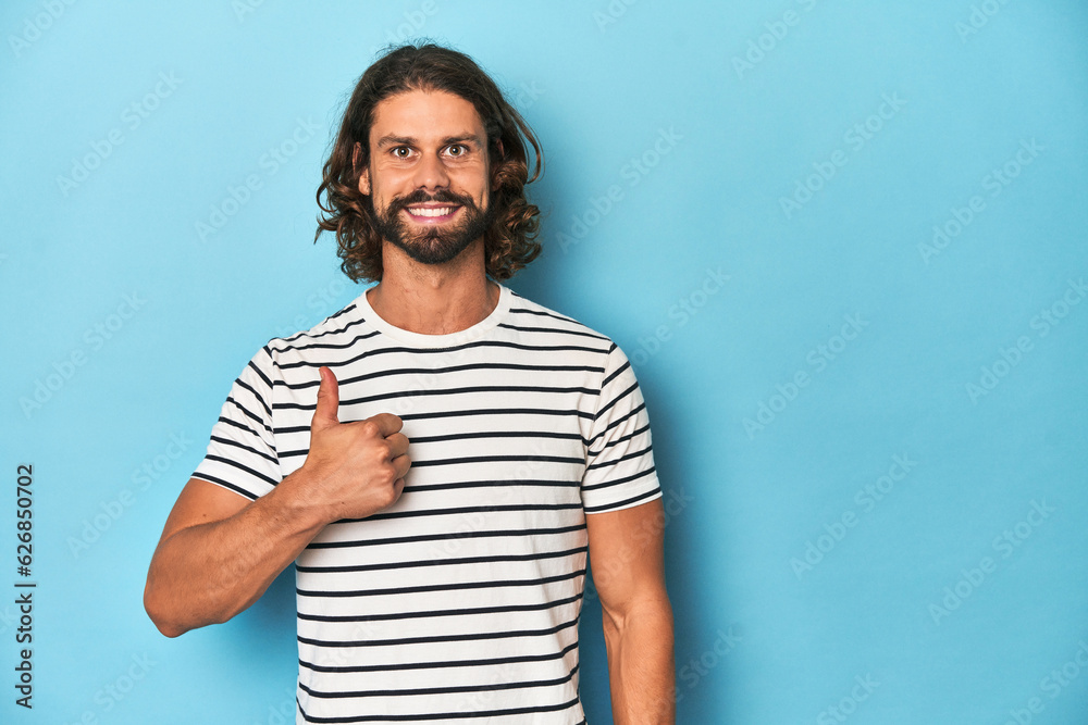 Bearded man in a striped shirt, blue backdrop smiling and raising thumb up Bearded man in a striped shirt, blue backdrop smiling and raising thumb up