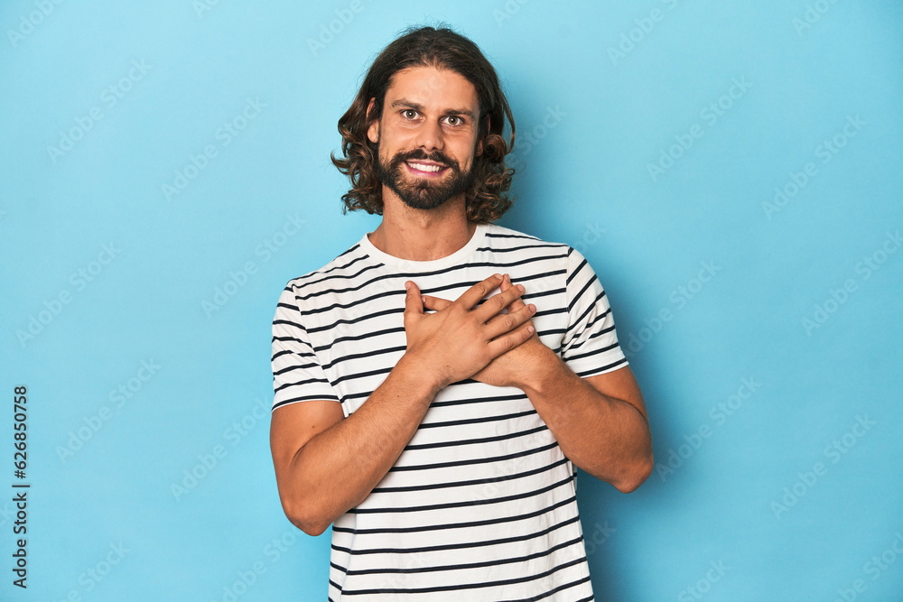 Bearded man in a striped shirt, blue backdrop has friendly expression, pressing palm to chest. Love concept. Bearded man in a striped shirt, blue backdrop has friendly expression, pressing palm to chest. Love concept.