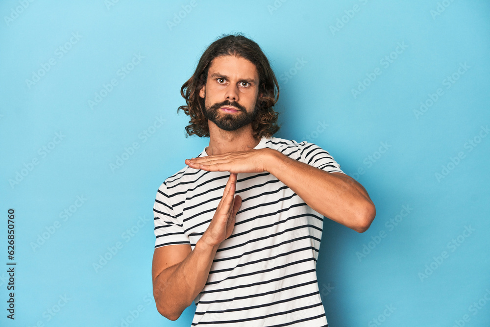 Bearded man in a striped shirt, blue backdrop showing a timeout gesture.