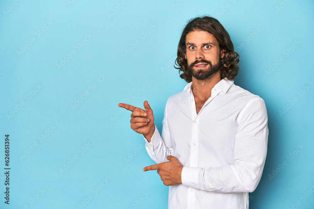 Bearded man in a white shirt, blue backdrop shocked pointing with index fingers to a copy space. Bearded man in a white shirt, blue backdrop shocked pointing with index fingers to a copy space.