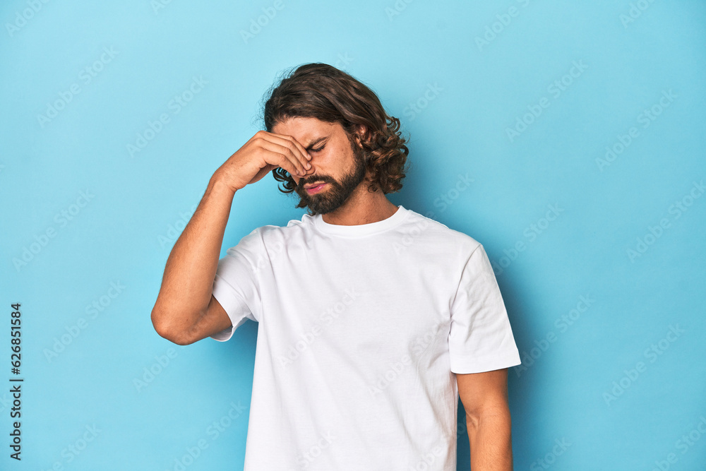 Bearded man in a white shirt, blue backdrop having a head ache, touching front of the face. Bearded man in a white shirt, blue backdrop having a head ache, touching front of the face.