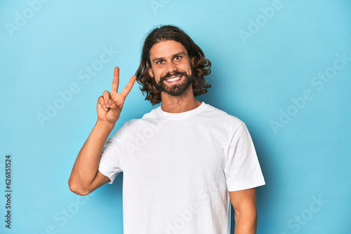 Bearded man in a white shirt, blue backdrop joyful and carefree showing a peace symbol with fingers.