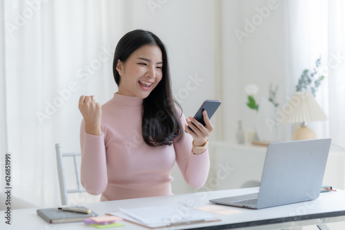 Happy young Asian woman raising hands with victory smiling happily with smart phone at home. Success, win, victory, triumph, congratulation, concept.