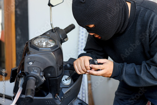 Motorcycle thief in black mask trying to unlock the ignition key of motorcycle with screwdriver. Street criminal, crime and people concept photo