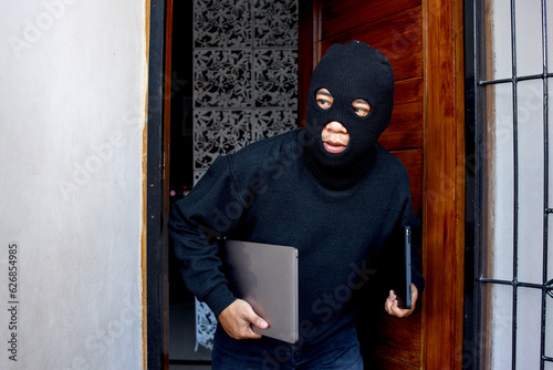 Sneaky and scared intruder wearing black balaclava mask stealing laptop from house. House intruder, housebreaking and burglary concept photo