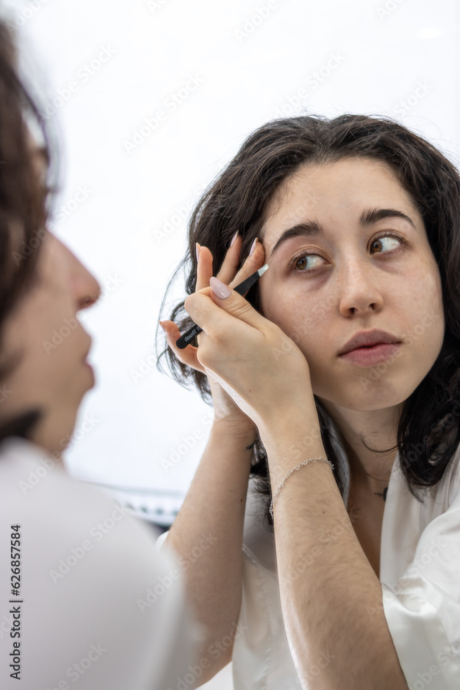 Attentive concentrated young latin female standing in bathroom at morning, doing her eyebrow contours sharp accurate using tweezer, plucking tweezing correcting thick brows, removing hairs Attentive concentrated young latin female standing in bathroom at morning, doing her eyebrow contours sharp accurate using tweezer, plucking tweezing correcting thick brows, removing hairs