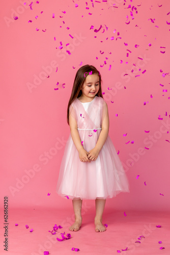 little girl in a pink dress catches confeti smiling happy on pink background, holiday concept. A child is celebrating a birthday on a pink background