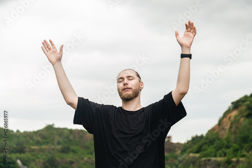 A young Christian man with a beard in the mountains in nature raises his hands to the sky, thanks God, praises.
