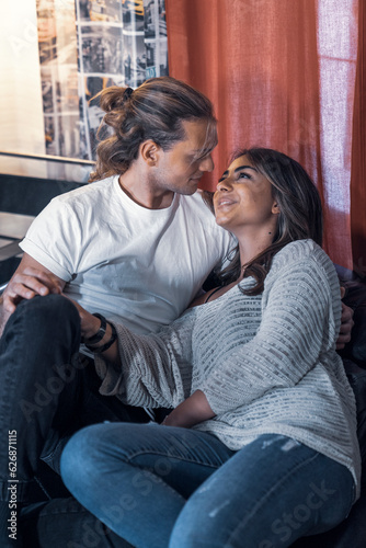 Inside house couple shooting sitting on sofa relaxing and looking at each other eyes smiling - Yellow and blue atmosphere tone light. Lovers shot talking in house studio red courtain