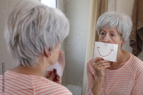 Sad senior woman holding a drawing with a smile in front of the mirror 