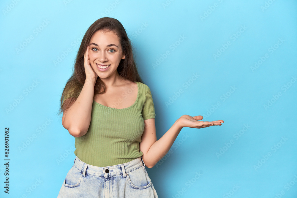 Young Caucasian woman in a green top on a blue backdrop holds copy space on a palm, keep hand over cheek. Amazed and delighted. Young Caucasian woman in a green top on a blue backdrop holds copy space on a palm, keep hand over cheek. Amazed and delighted.