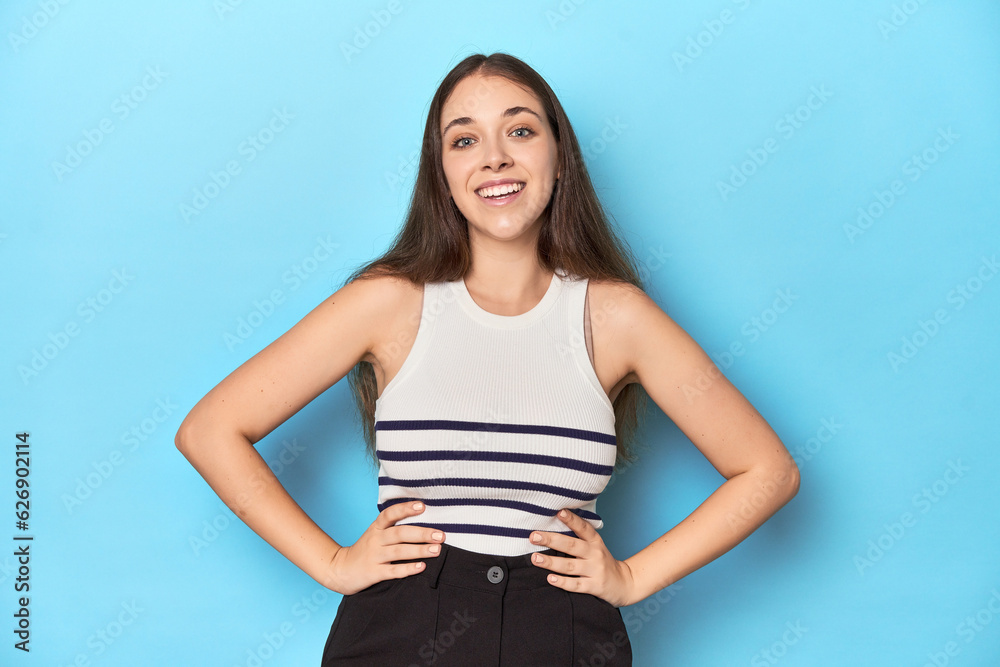Woman in a striped top posing on a blue studio backdrop confident keeping hands on hips. Woman in a striped top posing on a blue studio backdrop confident keeping hands on hips.