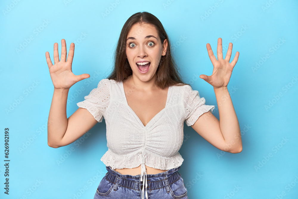 Stylish young woman in white blouse on a blue studio backdrop receiving a pleasant surprise, excited and raising hands.