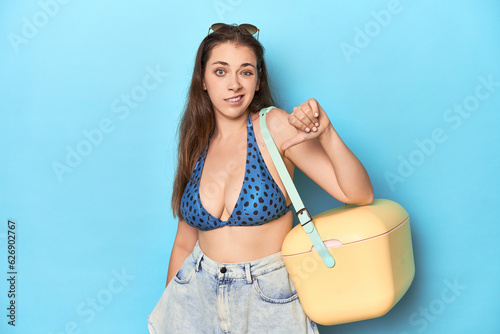 Woman in bikini with a portable beach cooler, blue studio showing a dislike gesture, thumbs down. Disagreement concept.