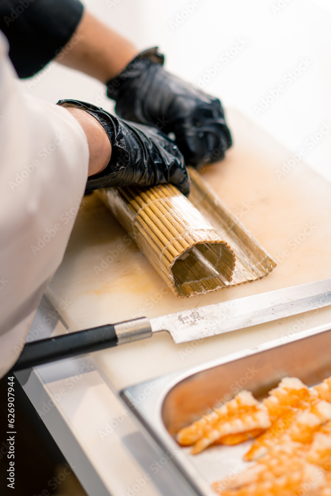 Close-up of a sushi maker in black gloves wrapping a california roll with a bamboo sushi mat in a professional kitchen 