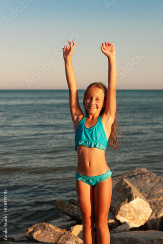 Beautiful cauxasian girl in a blue swimsuit with long hair at sunset on the seashore looks at the camera and smiles