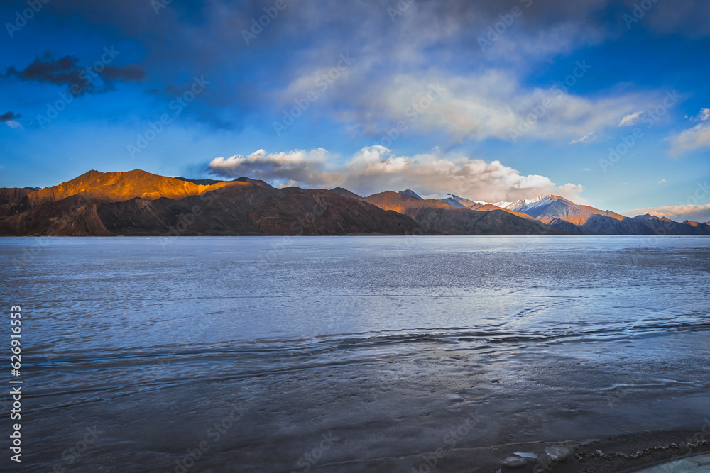 View of dark brown mountains capped with glistening snow in the distance, standing tall against the frozen beauty of Pangong Lake, the world's highest saltwater lake