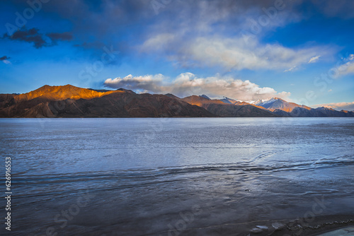 View of dark brown mountains capped with glistening snow in the distance, standing tall against the frozen beauty of Pangong Lake, the world's highest saltwater lake