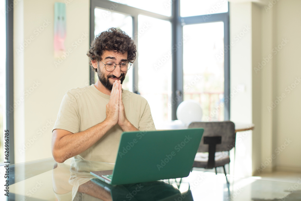 young adult bearded man with a laptop feeling worried, hopeful and religious, praying faithfully with palms pressed, begging forgiveness