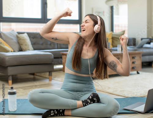 young adult woman practicing yoga feeling happy, positive and successful, celebrating victory, achievements or good luck