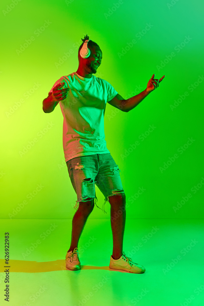 Full length image of dancing young man with dreadlocks in headphones with raising hand up over gradient colorful background in neon. Full length image of dancing young man with dreadlocks in headphones with raising hand up over gradient colorful background in neon.