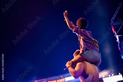 Rear view of man carrying his girlfriend on his shoulders during open air music concert at night.